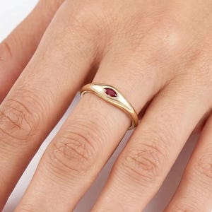 May include: A gold band ring featuring a single marquise-cut red gemstone set horizontally in the center. The ring has a smooth, polished finish and a tapered design that narrows toward the back, showcasing a clean and simple aesthetic.