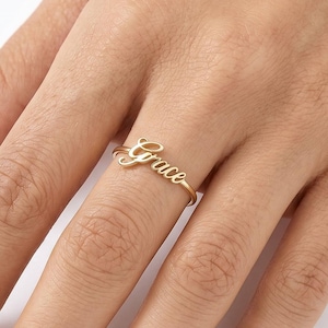 Op de afbeelding: Een goudkleurige ring met de naam Grace in een cursief lettertype. De ring heeft een dunne, gepolijste band die om de vinger sluit, met het naamplaatje gecentreerd op de bovenkant van de band, voorzien van een gladde, metalen afwerking.
