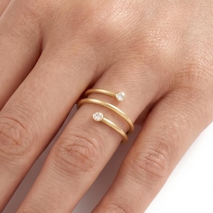 May include: A gold bypass ring featuring a spiral design that wraps around the finger. The ring is crafted from a smooth, matte-finish gold band and is accented with two small, round, clear white stones set at each open end of the spiral.