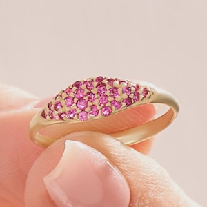 May include: A gold ring with a curved, oval-shaped top covered in small, bright pink gemstones. The ring is held between fingers against a soft pink background, showcasing the ring's design and color.