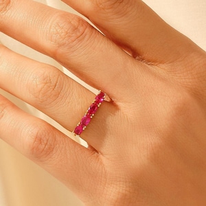 May include: A gold band ring featuring a row of five vibrant, oval-cut pink gemstones set in a prong setting. The ring is worn on a finger, showcasing the polished metal band and the consistent size and deep pink color of the five stones.