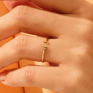 May include: A thin gold open ring featuring a small, delicate letter M at one end. The band is slender and polished, designed with an open gap that allows for slight adjustment. The metal has a bright, reflective finish that catches the light.