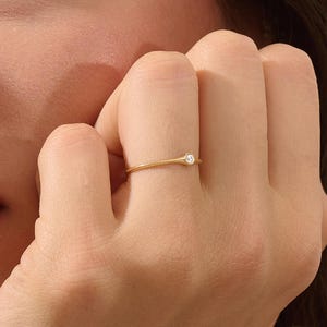 May include: A delicate gold ring with a small, round, clear gemstone. The ring is simple and elegant, with a thin band. The ring is on a person's finger.
