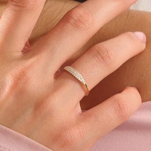 May include: A gold ring featuring a curved, tapered band set with a dense row of small, clear, sparkling stones across the top surface. The band is thin and polished, showcasing a bright metallic finish that contrasts with the embedded stones.