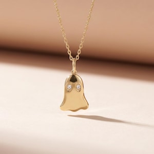 May include: A gold-colored ghost pendant necklace with a delicate chain. The ghost pendant has two small, round, clear gemstones for eyes. The necklace is displayed against a soft, neutral background.