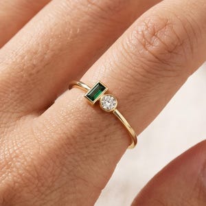 Könnte beinhalten: Ein Goldring mit einem kleinen, rechteckig geschliffenen grünen Edelstein im Smaragdschliff und einem einzelnen runden, klaren Edelstein im Brillantschliff, die nebeneinander auf einem dünnen, polierten Goldring sitzen.