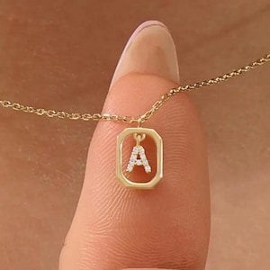 May include: A fine gold necklace with a pendant displaying the letter "A" within a rectangular setting. The pendant is embellished with tiny, glittering stones. The necklace is shown on a person's finger.