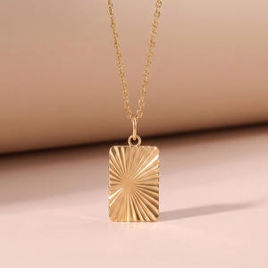 May include: A gold-colored necklace with a rectangular pendant. The pendant has a sunburst design and hangs from a delicate chain. The background is a soft, neutral color.