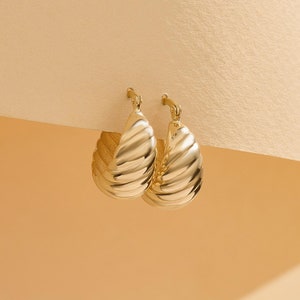May include: A pair of gold hoop earrings featuring a thick, croissant-inspired ribbed texture. The earrings have a polished, reflective finish and a hinged post closure mechanism, showcasing a rounded, chunky silhouette with a curved design.