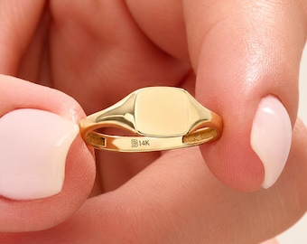 May include: A polished 14K yellow gold signet ring featuring a smooth, flat, square-shaped face. The interior of the band is clearly stamped with the text 14K, indicating the gold purity of this solid metal jewelry piece.