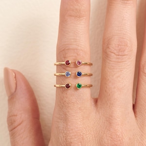 Puede incluir: Tres anillos abiertos de oro apilados en un dedo, cada uno con dos piedras preciosas engastadas en bisel. El anillo superior tiene una piedra roja y una rosa, el del medio tiene dos piedras azules y el anillo inferior tiene una piedra roja oscura y una verde.
