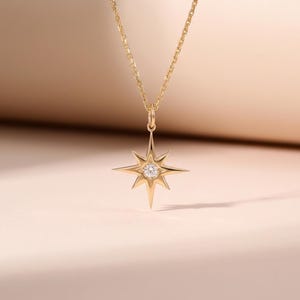 May include: A gold-colored necklace featuring a starburst pendant with a central, round, clear gemstone. The pendant has pointed rays extending outwards. The chain is delicate and gold-toned, with a simple clasp.