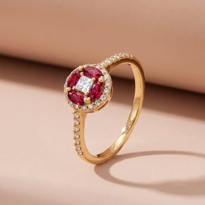 May include: A gold ring featuring a central square white gemstone surrounded by a halo of smaller white gemstones and oval-cut red gemstones. The ring's band is also adorned with small white gemstones. The ring is set against a soft pink background.