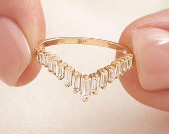 May include: A gold V-shaped ring featuring a row of clear baguette-cut stones set along the top edge. The band is stamped with the text 14K on the inner surface. The design follows a chevron silhouette with multiple rectangular stones.