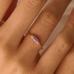 May include: A gold ring with a light purple oval-shaped gemstone. The ring has a thin band and a decorative setting around the gemstone. The ring is on a finger.