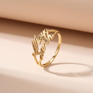 May include: A gold-colored ring with the name "Maya Sarah" in a cursive font. The ring is set against a soft, neutral background, highlighting the jewelry's design and color. The ring is a personalized accessory.