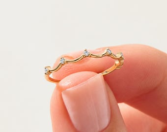 May include: A thin gold ring featuring a delicate, wavy band design. The top of the band is accented with five small, clear, round-cut stones set at regular intervals along the curved, crown-like silhouette of the metal band.