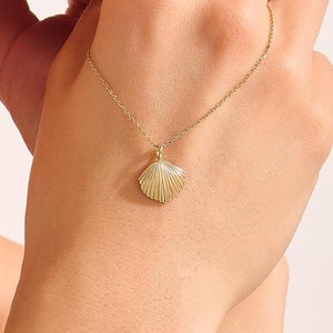 May include: A delicate gold necklace featuring a seashell pendant. The pendant is intricately detailed with a ridged texture, suspended from a fine chain. The necklace is displayed on a person's hand, showcasing its elegant design.