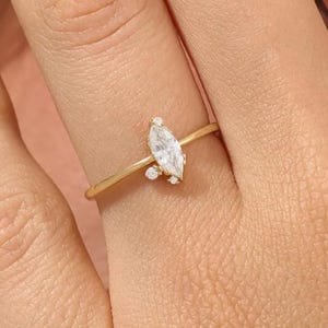 May include: A gold ring featuring a marquise-cut diamond center stone and small accent diamonds. The ring has a delicate band and is set with a cluster of small diamonds on one side of the center stone.