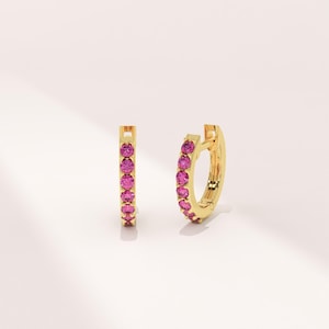 Könnte beinhalten: Ein Paar goldene Creolen, jeweils mit einer Reihe leuchtend rosa Edelsteinen besetzt. Die Ohrringe werden vor einem weichen, neutralen Hintergrund präsentiert, der ihre polierte, reflektierende Oberfläche und das Funkeln der Edelsteine hervorhebt.