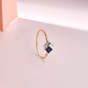 May include: A gold ring featuring a cluster of gemstones. The ring has a delicate band and is adorned with a dark blue square-cut stone, a light blue stone, and a clear stone. Small, clear stones are set on the band.