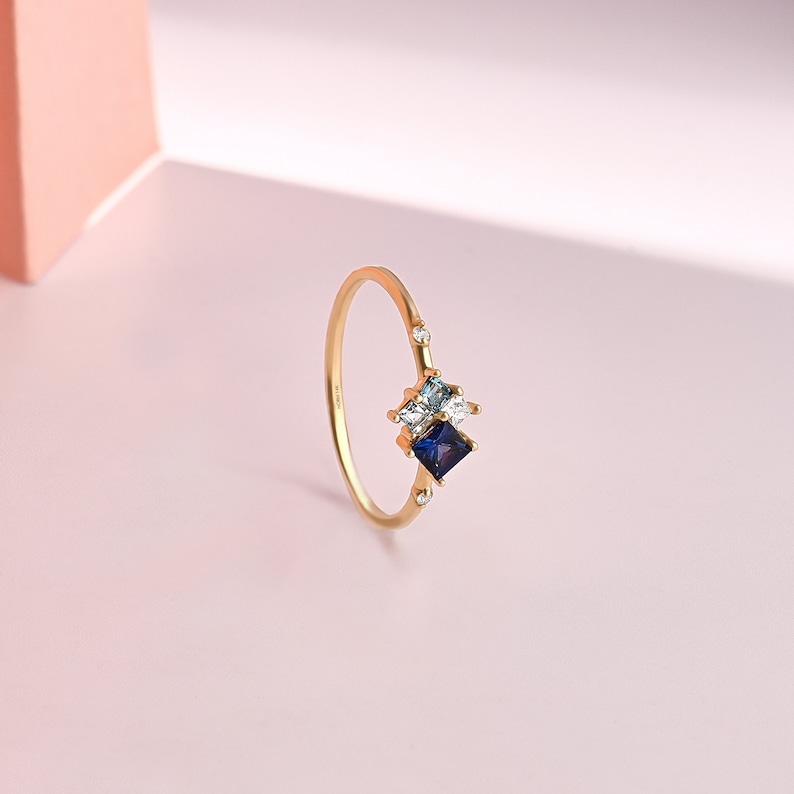 May include: A gold ring featuring a cluster of gemstones. The ring has a delicate band and is adorned with a dark blue square-cut stone, a light blue stone, and a clear stone. Small, clear stones are set on the band.