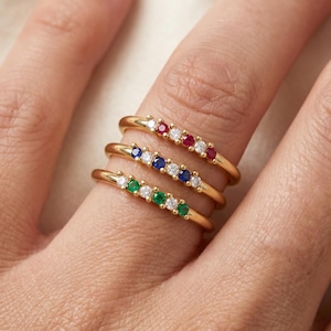 May include: Three gold rings stacked on a finger, each featuring a row of five alternating round gemstones. The top ring has red and clear stones, the middle ring has blue and clear stones, and the bottom ring has green and clear stones.