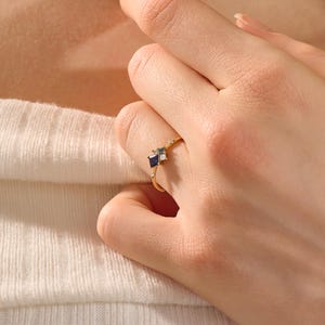 May include: A gold ring featuring a cluster of square and rectangular gemstones in shades of blue and white. The ring has a delicate band and is adorned with small, sparkling accents. The ring is on a finger.