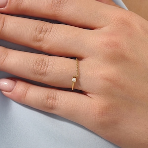 May include: A delicate gold ring featuring a thin band that transitions into a fine chain segment, accented by a single small, round, clear gemstone set in a square-shaped prong setting, positioned on the finger between the knuckle and the base.
