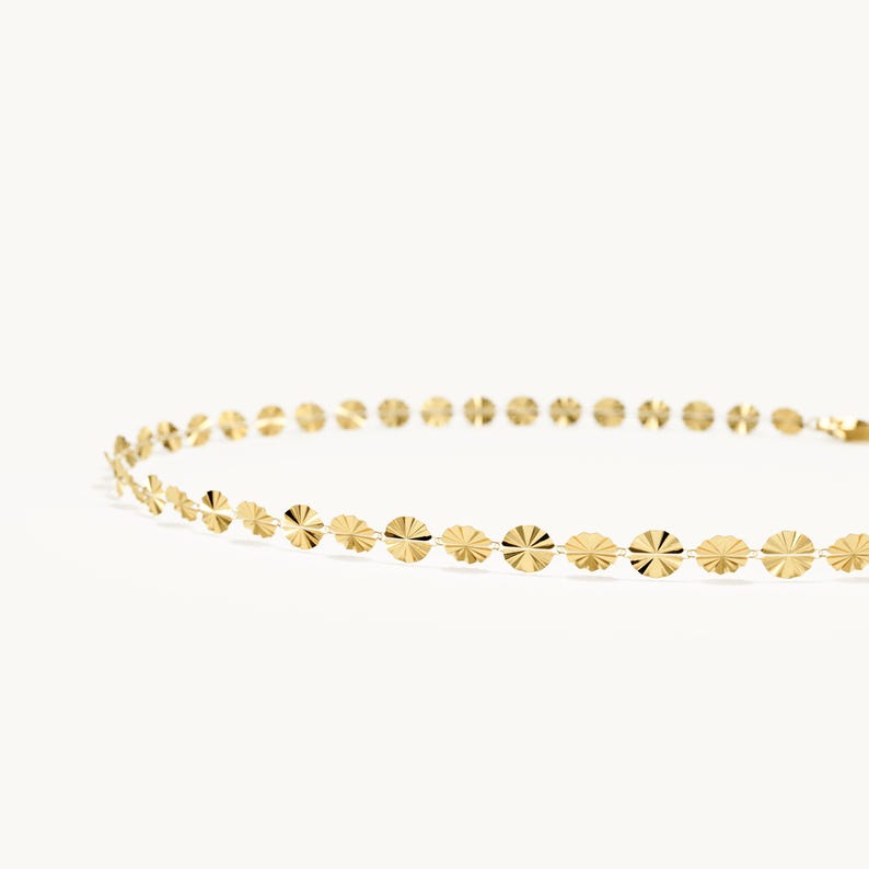 May include: A gold chain necklace featuring a series of small, flat, circular discs with a sunburst-like textured pattern. The discs are linked together in a continuous row, creating a delicate and reflective metallic jewelry piece.