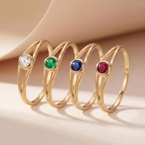 May include: Four gold rings with different colored gemstones: a clear diamond, a green emerald, a blue sapphire, and a red ruby. Each ring has a unique gemstone set in a gold band with a split shank design. The rings are displayed on a light pink background.