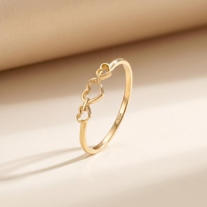 May include: A delicate gold ring featuring three connected heart-shaped designs. The ring is made of polished gold and has a slender band. The inscription "14K" is visible on the ring's band, indicating the gold's purity.