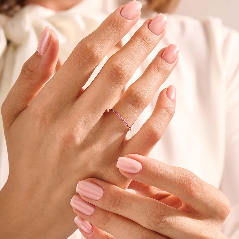 Puede incluir: Un delicado anillo de oro adornado con una l&iacute;nea continua de peque&ntilde;as y vibrantes piedras preciosas rojas. El anillo se muestra en una mano con u&ntilde;as cuidadas, sobre un fondo suave y neutro. El dise&ntilde;o del anillo es simple pero elegante.