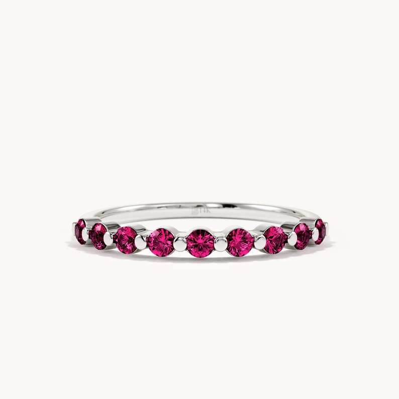May include: A silver-toned ring featuring a series of round, vibrant pink gemstones set in a row. The band is thin and metallic, with small silver beads positioned between each of the circular pink stones, creating a continuous, symmetrical design.