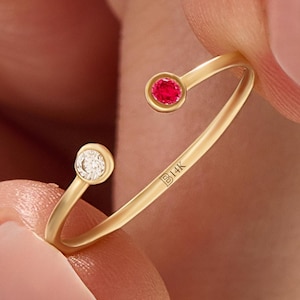 Könnte beinhalten: Ein goldfarbener offener Ring mit einem kleinen, runden, roten Edelstein und einem kleinen, runden, klaren Edelstein. Der Ring ist mit "B14K" markiert. Der Ring hat ein minimalistisches Design.