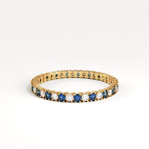 May include: A gold band ring with alternating blue and white gemstones. The ring is set against a white background, highlighting the sparkle of the gemstones. The ring is a classic eternity style.