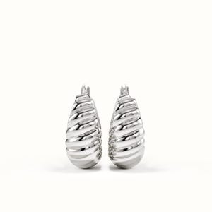 May include: A pair of silver-toned hoop earrings featuring a thick, rounded, and ribbed croissant-inspired texture. The earrings have a polished, reflective metallic finish with a hinged post closure at the top, showcasing a bold, sculptural design.