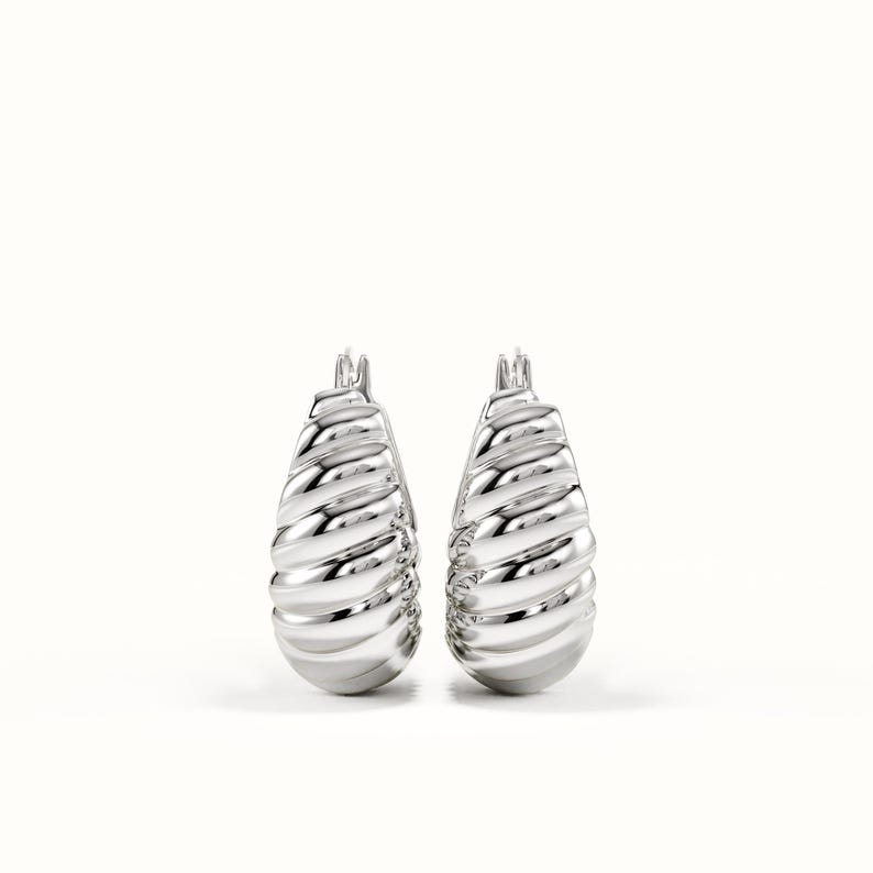 May include: A pair of silver-toned hoop earrings featuring a thick, rounded, and ribbed croissant-inspired texture. The earrings have a polished, reflective metallic finish with a hinged post closure at the top, showcasing a bold, sculptural design.