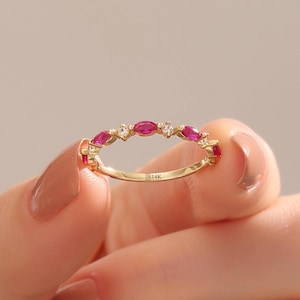 May include: A gold ring featuring a series of marquise-cut pink gemstones and small, round, clear gemstones. The ring is held by a person. The band is stamped with "B14K".