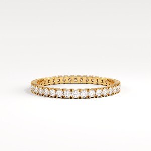 May include: A gold eternity ring with a continuous row of round, clear gemstones. The ring is set in a yellow gold band and sits on a white background. The gemstones are evenly spaced around the entire band.