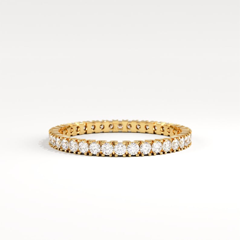 May include: A gold eternity ring with a continuous row of round, clear gemstones. The ring is set in a yellow gold band and sits on a white background. The gemstones are evenly spaced around the entire band.