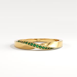 May include: A gold-colored ring with a twisted design. The ring is adorned with a row of small, round, green gemstones. The ring is set against a white background, highlighting its elegant and modern style. The ring is marked with "14K".