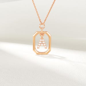 May include: A rose gold necklace with a pendant shaped like the letter "A", embellished with small, sparkling stones. The pendant is set within a rectangular frame, suspended from a delicate chain. The necklace is displayed against a white backdrop.