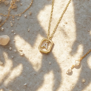 May include: A gold-coloured necklace with a pendant featuring the letter "A" encrusted with small, clear gemstones. The pendant is set within a square frame. Other gold necklaces and small stones are scattered around the main necklace.