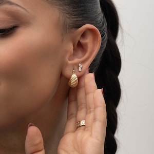 May include: A gold croissant-style hoop earring with a ribbed texture, paired with a small stud earring featuring two clear baguette-cut stones. A gold band ring with a repeating sun and moon motif is worn on the finger near the earlobe.