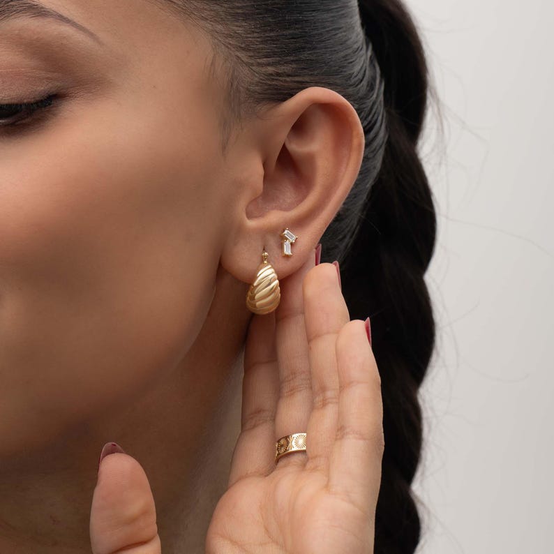May include: A gold croissant-style hoop earring with a ribbed texture, paired with a small stud earring featuring two clear baguette-cut stones. A gold band ring with a repeating sun and moon motif is worn on the finger near the earlobe.