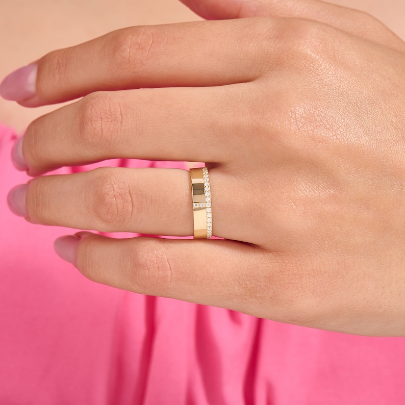 May include: A gold ring with a rectangular design and a row of small, clear stones. The ring is on a hand with pink nail polish, set against a pink background.