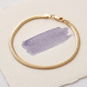 May include: A gold-toned herringbone chain bracelet featuring a flat, flexible link design that creates a continuous, woven pattern. The bracelet is secured with a small, functional lobster claw clasp at one end, allowing for easy fastening.