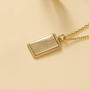May include: A gold-toned necklace featuring a rectangular pendant with a smooth, polished centre surface and a textured, rope-style border. The pendant is attached to a delicate gold-toned chain by a small circular jump ring.