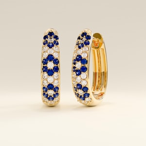 Könnte beinhalten: Ein Paar goldene Creolen. Jeder Ohrring ist mit einem Muster aus blauen und weißen Edelsteinen verziert. Die Edelsteine sind in einem floralen Design angeordnet. Die Ohrringe haben eine polierte, reflektierende Oberfläche.